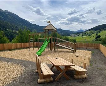 Ein Spielplatz mit einer Rutsche und Klettergerüst in einer naturnahen Umgebung. Im Hintergrund sind Berge und ein blauer Himmel zu sehen.