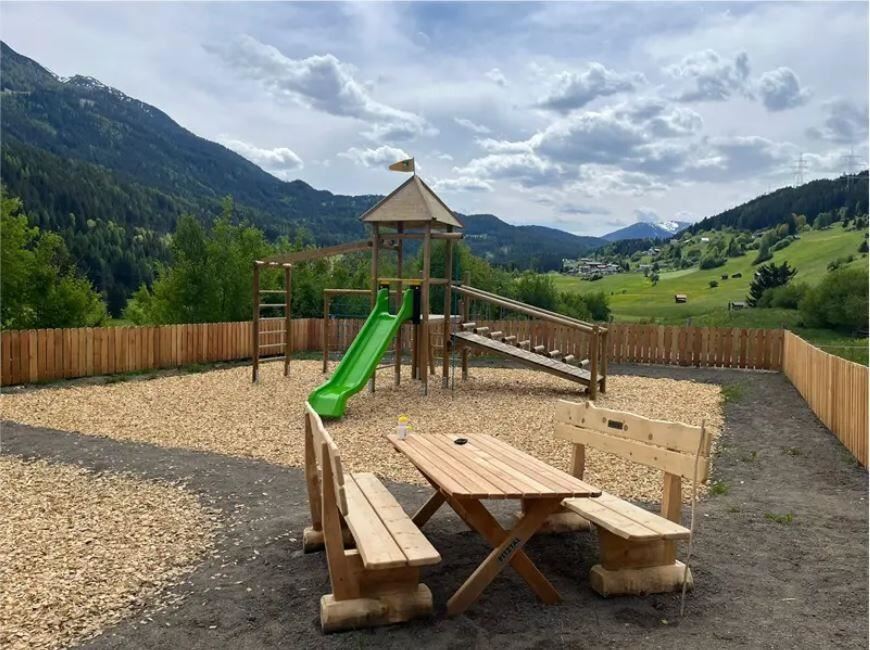 Ein Spielplatz mit einer Rutsche und Klettergerüst in einer naturnahen Umgebung. Im Hintergrund sind Berge und ein blauer Himmel zu sehen.