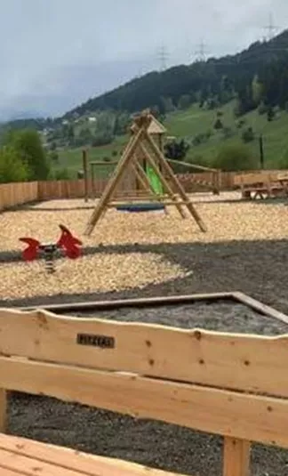 Ein Spielplatz in einer grünen Landschaft, umgeben von Bäumen und Hügeln. Es gibt Holzspielgeräte und eine Sitzbank im Vordergrund.
