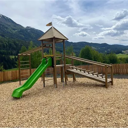 Ein Spielplatz mit einer Rutsche und einer Kletterstation in einer schönen, bergigen Landschaft. Die Umgebung ist von Bäumen umgeben und die Wolken am Himmel verleihen eine freundliche Atmosphäre.