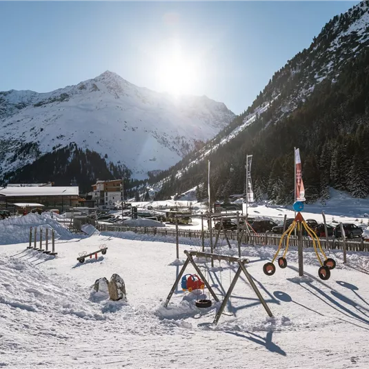 Ein verschneites Bergdorf mit Spielplatz im Vordergrund. Im Hintergrund sind hohe Berge und ein klarer Himmel zu sehen.
