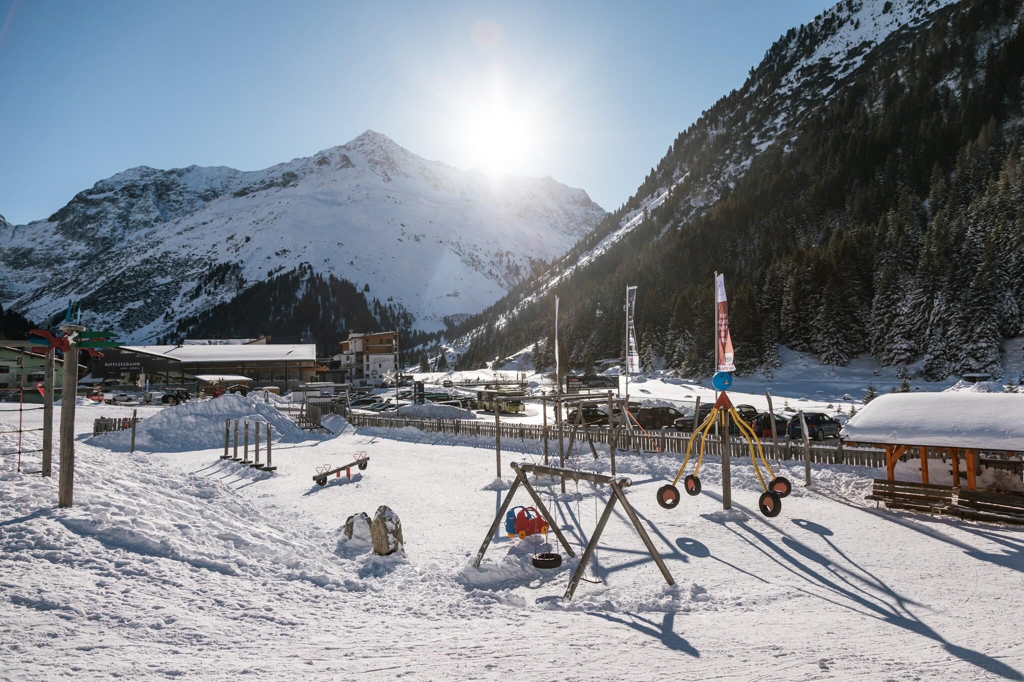 Ein verschneites Bergdorf mit Spielplatz im Vordergrund. Im Hintergrund sind hohe Berge und ein klarer Himmel zu sehen.