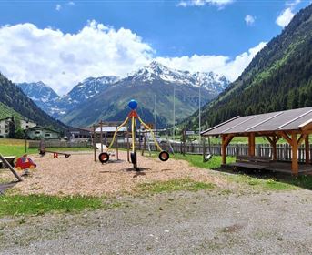 A playground in the mountains with swinging equipment and a covered seating area. In the background, green hills and snow-capped mountains can be seen.