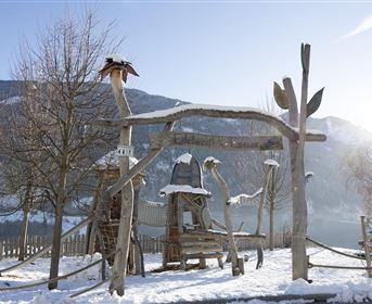 Eine Winterlandschaft mit schneebedecktem Boden. Im Vordergrund steht ein kreativer Spielplatz aus Holz mit verschiedenen Elementen.