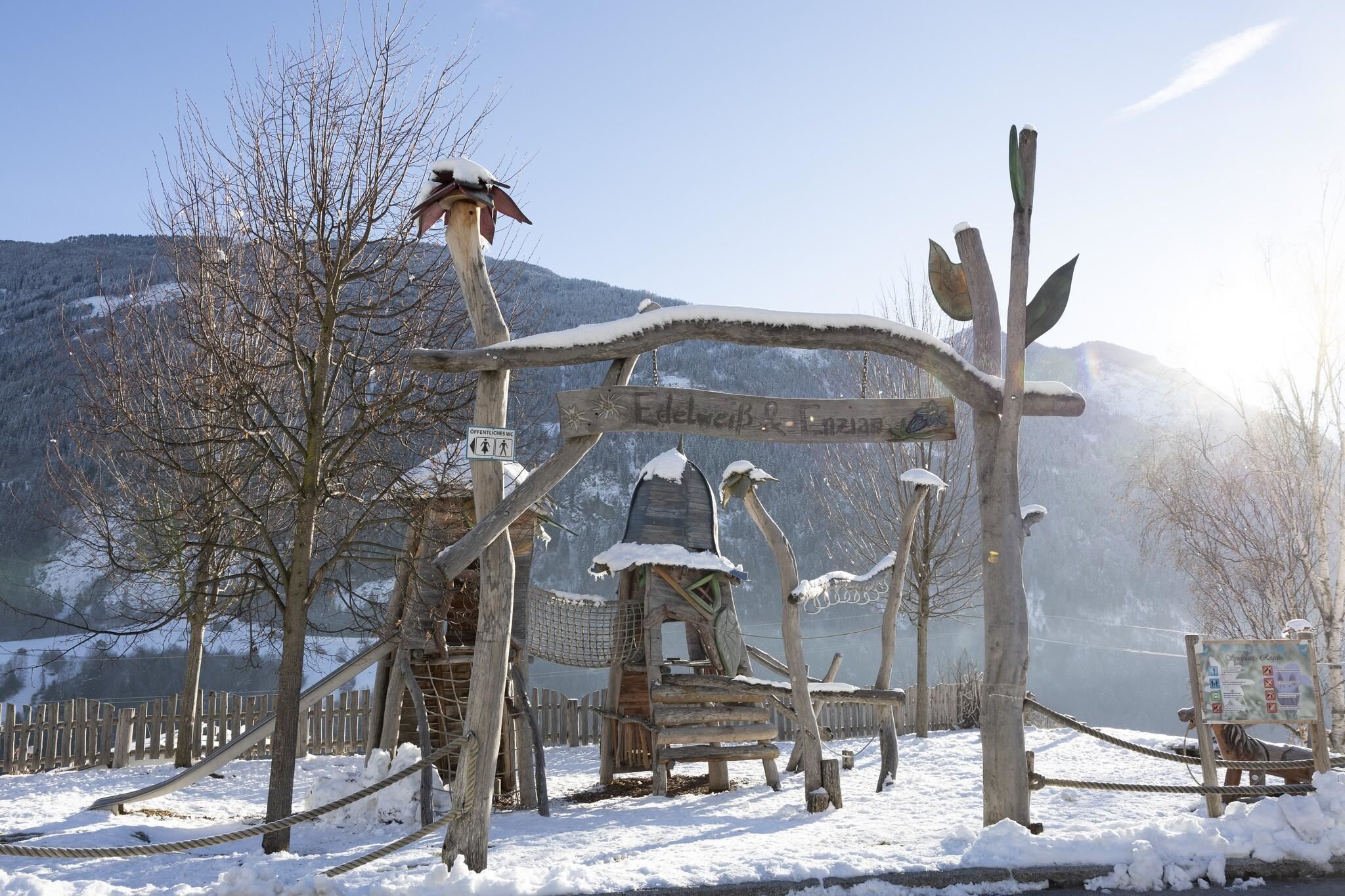 Eine Winterlandschaft mit schneebedecktem Boden. Im Vordergrund steht ein kreativer Spielplatz aus Holz mit verschiedenen Elementen.