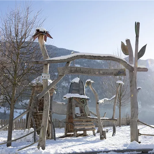 Eine Winterlandschaft mit schneebedecktem Boden. Im Vordergrund steht ein kreativer Spielplatz aus Holz mit verschiedenen Elementen.
