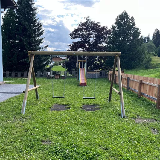 Ein schöner Spielplatz mit einer Schaukel und einer Rutsche. Im Hintergrund sind Bäume und eine Wiese zu sehen.