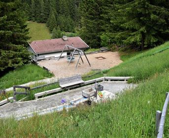 Ein Spielplatz in der Natur mit einer Schaukel und einem gemütlichen Holzhaus im Hintergrund. Umgeben von hohen Bäumen und grünen Wiesen.