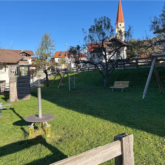 A playground with green meadows, a swing, and a climbing frame. In the background, a church tower and some houses can be seen.