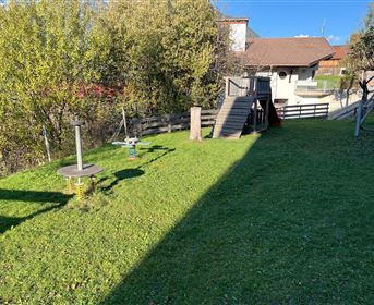 A well-maintained garden with green grass and a playground. In the background, a house can be seen, along with some trees and a blue sky.