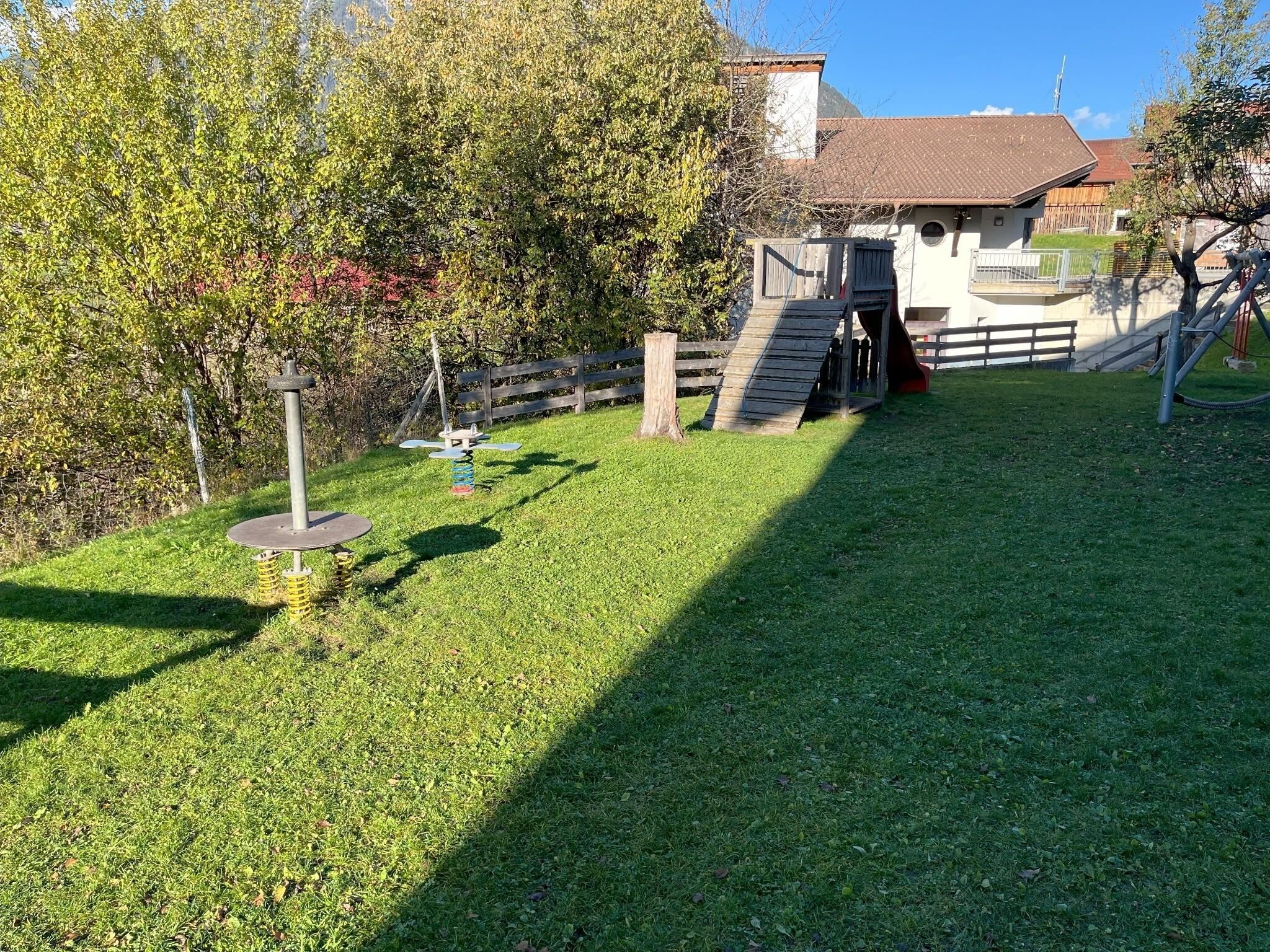 A well-kept garden with green grass and trees. In the background, there is a building with a wooden terrace and a slide.