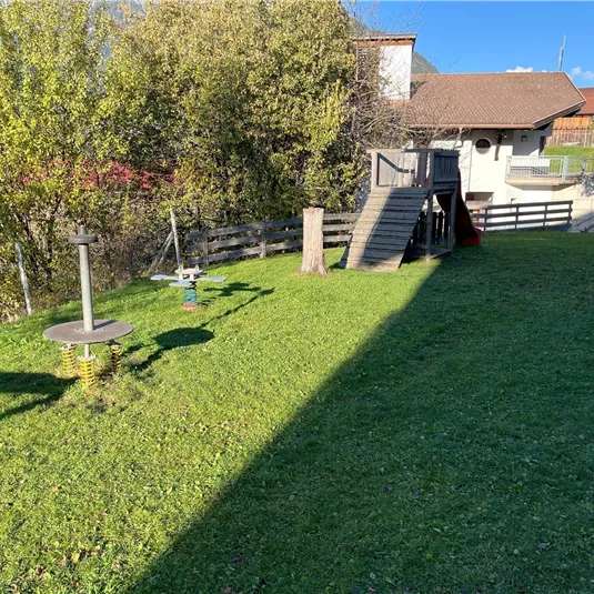 A well-kept garden with green grass and trees. In the background, there is a building with a wooden terrace and a slide.