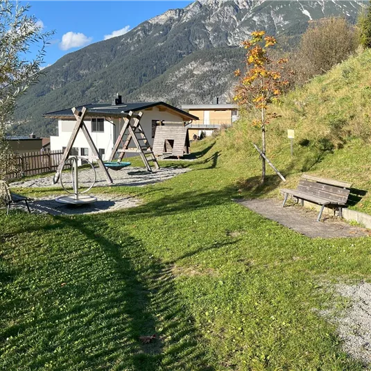 A playground with a swing and a bench, surrounded by green meadow. In the background, there are mountains and a modern house.