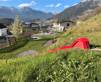 A playground with a red slide and swings, surrounded by green meadows. In the background, mountains and some houses can be seen.