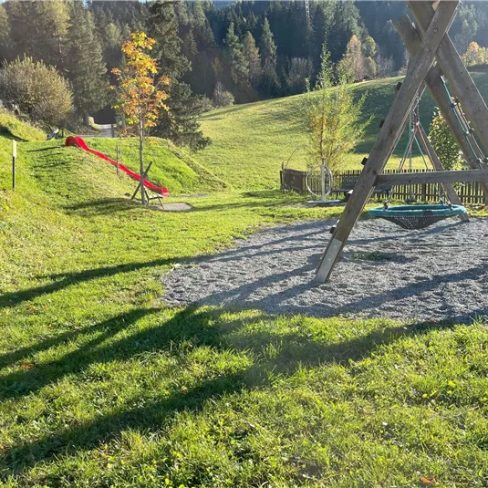 A playground with a slide and a seesaw is located on a green meadow. In the background, forests and gentle hills can be seen.