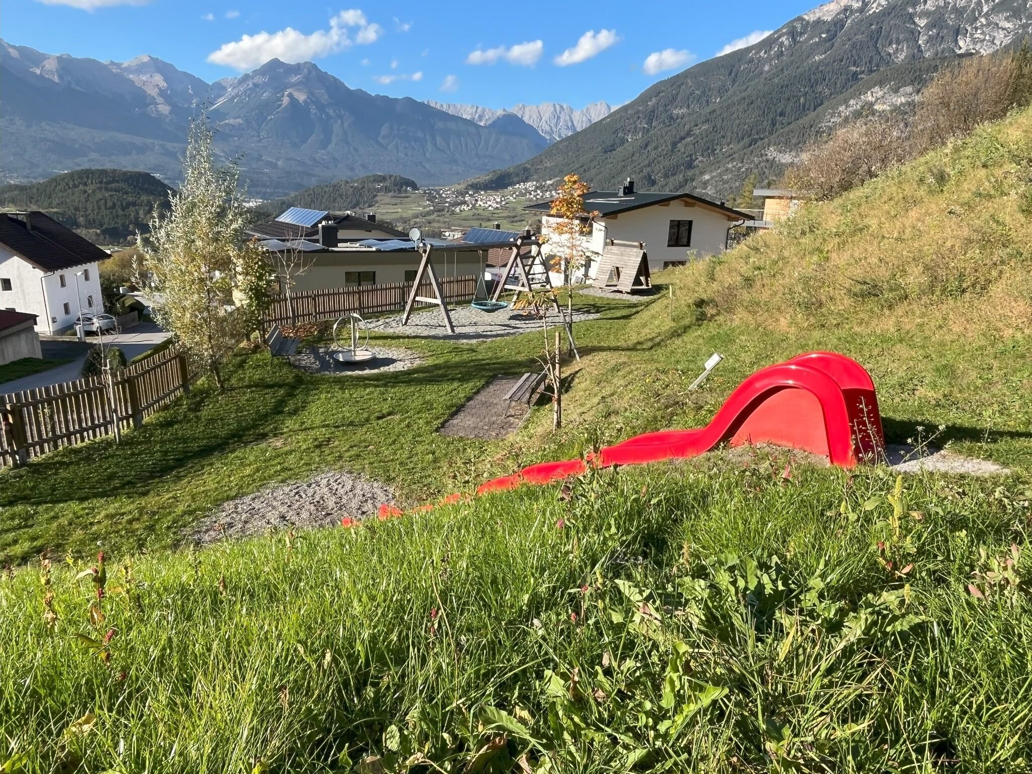 A playground in a picturesque landscape with mountains in the background. The red slide is in the foreground, surrounded by green grass and playground equipment.