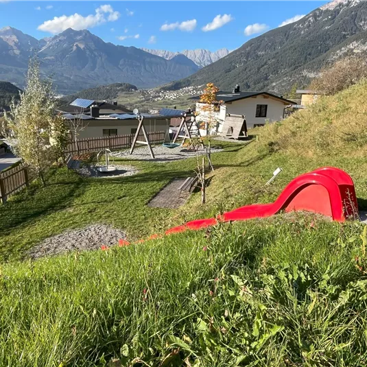 A playground in a picturesque landscape with mountains in the background. The red slide is in the foreground, surrounded by green grass and playground equipment.