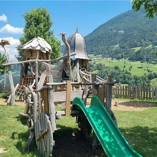 Ein Spielplatz mit einem Holzspielgerät und einer Rutsche. Im Hintergrund sind grüne Wiesen und Berge zu sehen.