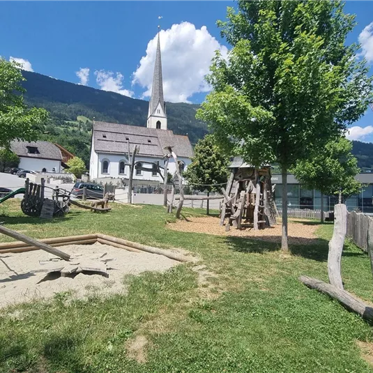 Ein Spielplatz mit Sandkasten und Spielgeräten in einer grünen Umgebung. Im Hintergrund sind ein Gebäude und ein Kirchturm vor einem Bergpanorama zu sehen.