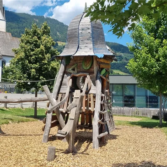 Ein Spielplatz mit einem Holzturm und einer Rutsche. Im Hintergrund sind Bäume und ein modernes Gebäude zu sehen.