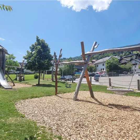 Ein Spielplatz mit einer Rutsche und Schaukeln, umgeben von grünem Rasen. Im Hintergrund sind Häuser und Bäume sichtbar.