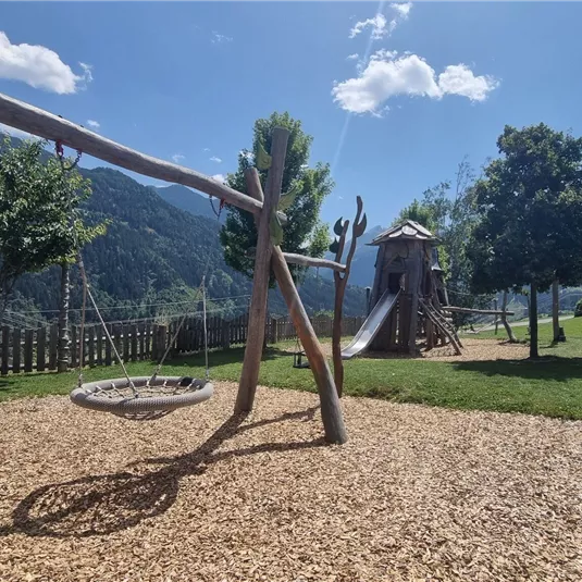Ein Spielplatz in der Natur mit Schaukeln, einem Rutschenturm und viel grüner Wiese. Im Hintergrund sind die Berge zu sehen und der Himmel ist klar und blau.