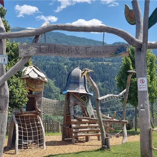 Ein Spielplatz mit einem großen Holzschild und bunten Spielgeräten. Im Hintergrund sieht man eine grüne Landschaft und Berge.
