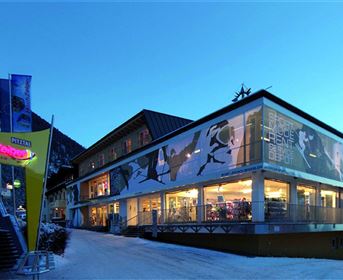 Ein modernes Gebäude in den Bergen, umgeben von Schnee. Die Fassade ist mit sportlichen Bildern dekoriert und es ist Abenddämmerung.