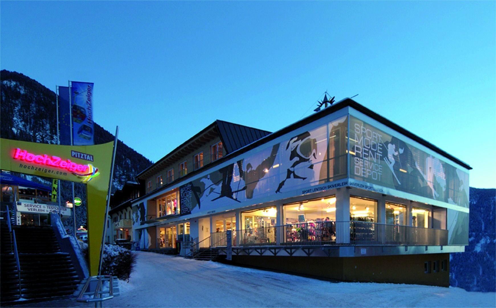 Ein modernes Gebäude mit großen Fenstern, das in den Bergen liegt. Es ist abends, und die Umgebung ist in sanftem Licht beleuchtet.