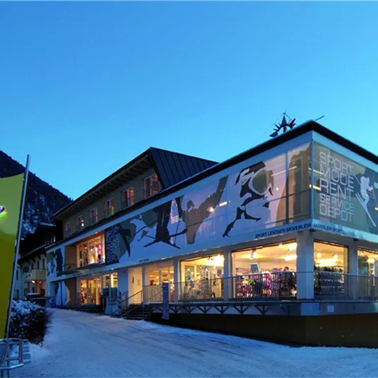 Ein modernes Gebäude mit großen Fenstern, das in den Bergen liegt. Es ist abends, und die Umgebung ist in sanftem Licht beleuchtet.