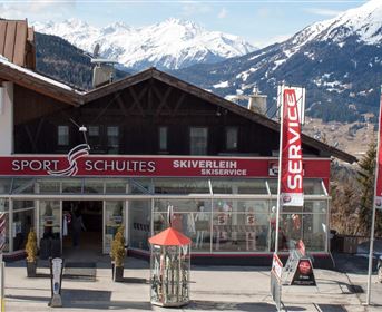 Ein Skiverleih mit dem Namen "Sport Schultes" in den Bergen. Im Hintergrund sind schneebedeckte Berge und ein blauer Himmel zu sehen.