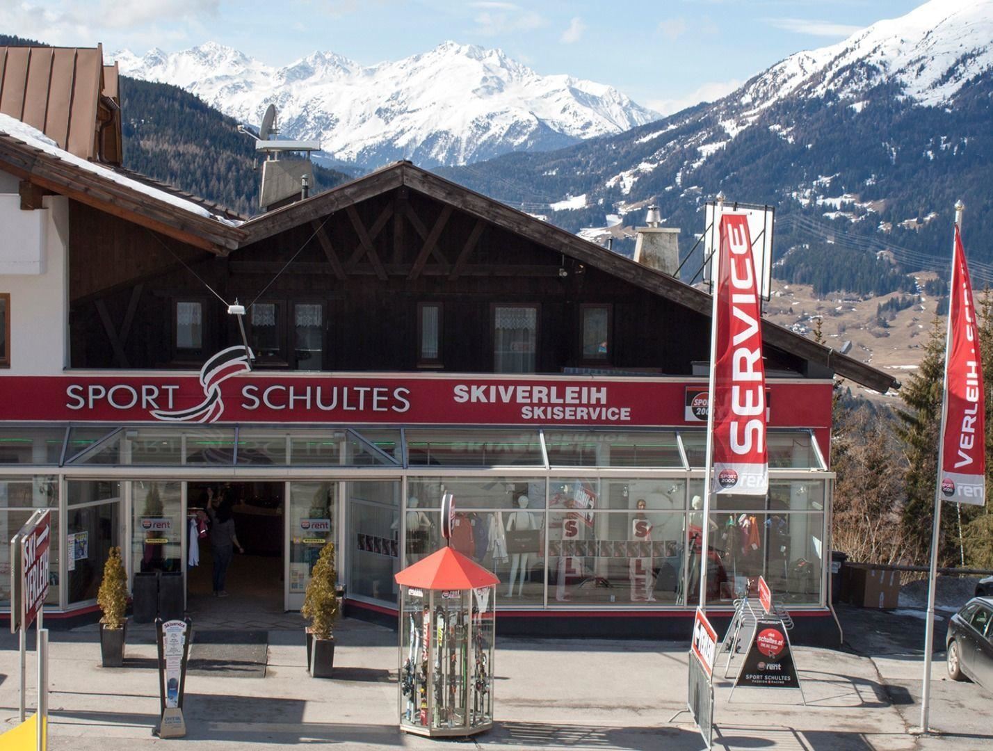 Ein Skiverleih mit dem Namen "Sport Schultes" in den Bergen. Im Hintergrund sind schneebedeckte Berge und ein blauer Himmel zu sehen.