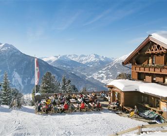 A cozy mountain house in the snow with guests sitting on the terrace. In the background, there are snow-covered mountains and a blue sky.