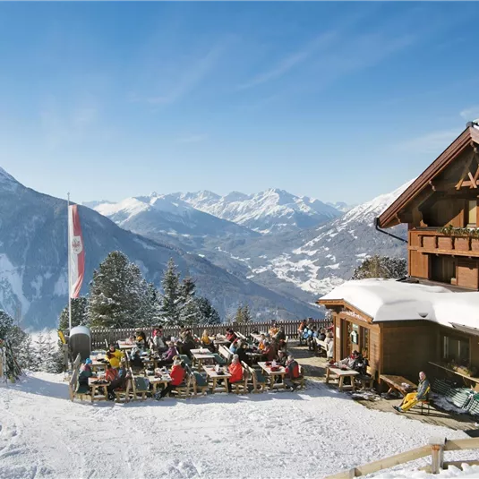 Ein gemütliches Berghaus im Schnee mit Gästen, die auf der Terrasse sitzen. Im Hintergrund sind schneebedeckte Berge und ein blauer Himmel zu sehen.