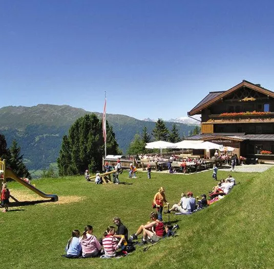 A cozy mountain hut with a sun terrace in a picturesque mountain landscape. In the foreground, children are playing on a playground, while families relax in the grass.