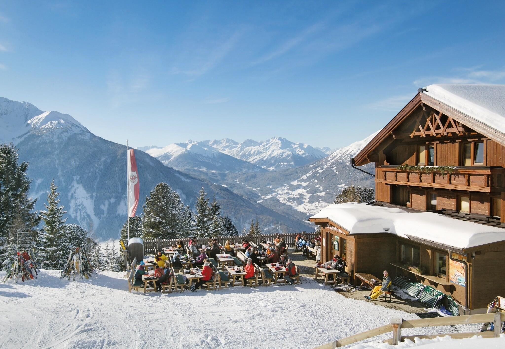 A picturesque mountain cabin in the snow with guests outside. In the background, majestic mountains rise under a clear blue sky.