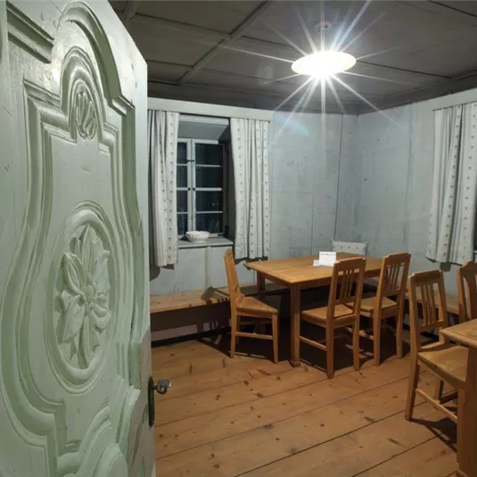 Ein gemütlicher Raum mit Holztisch und Stühlen. Helle Fenster lassen Licht herein und schaffen eine einladende Atmosphäre.