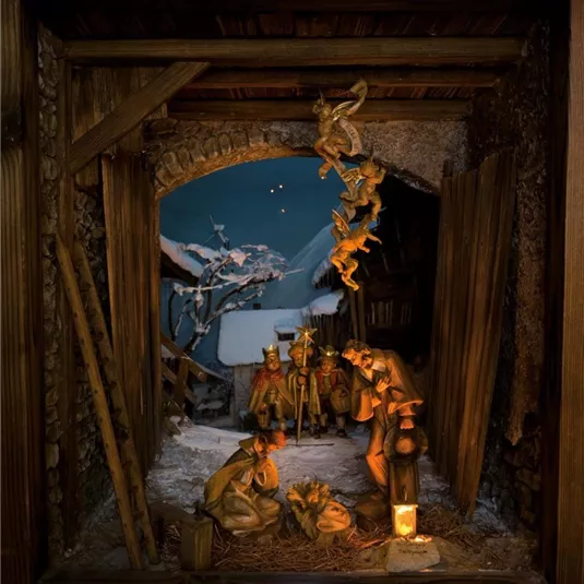 Eine traditionelle Krippe mit Figuren, die die Geburt Jesu darstellen. Im Hintergrund sind schneebedeckte Landschaften und ein klarer Sternenhimmel zu sehen.