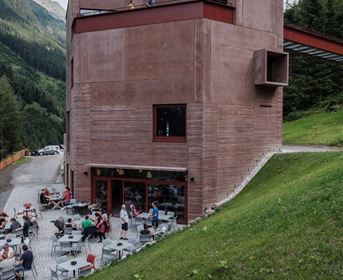 Ein modernes Gebäude in einer bergigen Umgebung mit einem gemütlichen Außenbereich. Die Terrasse ist voller Menschen, die entspannen und die Aussicht genießen.