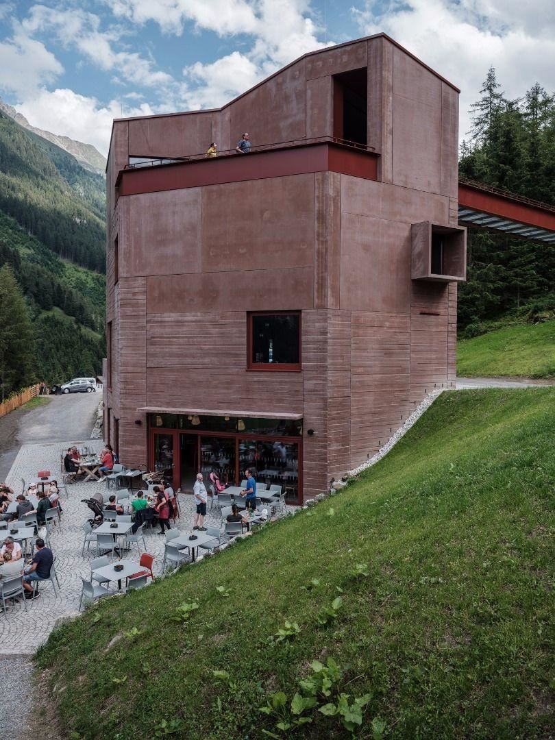 A modern building in the mountains with a terrace area. Many people are sitting outside and enjoying their leisure time.