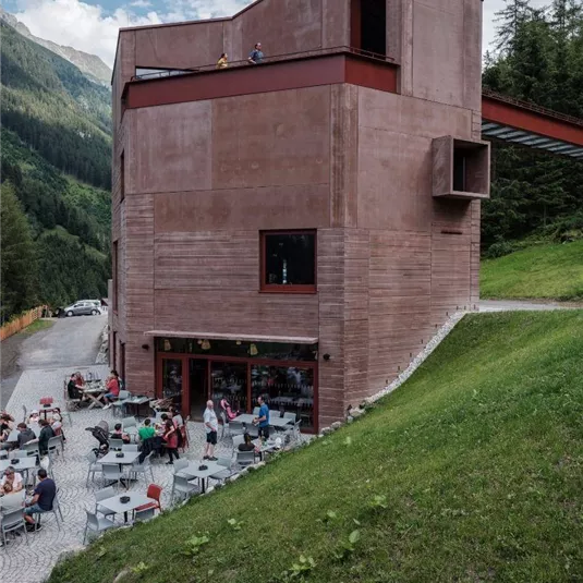 A modern building in the mountains with a terrace area. Many people are sitting outside and enjoying their leisure time.