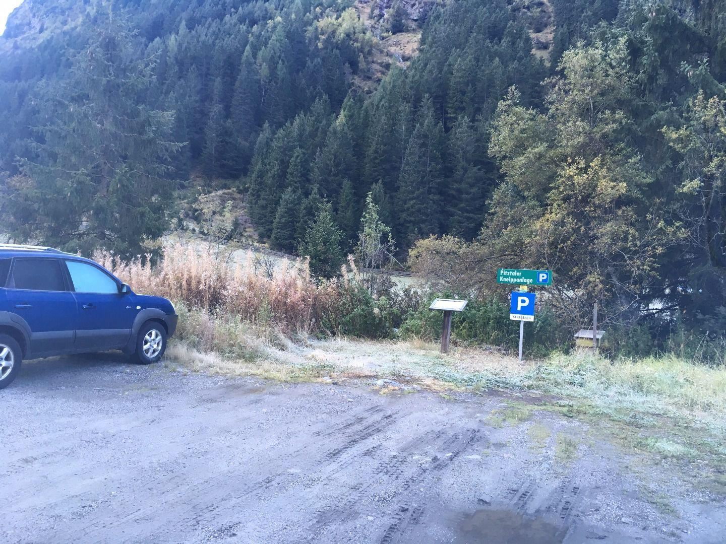 Ein Parkplatz in der Natur, umgeben von Bäumen und hoher Vegetation. Im Vordergrund steht ein blauer SUV auf einem unbefestigten Weg.
