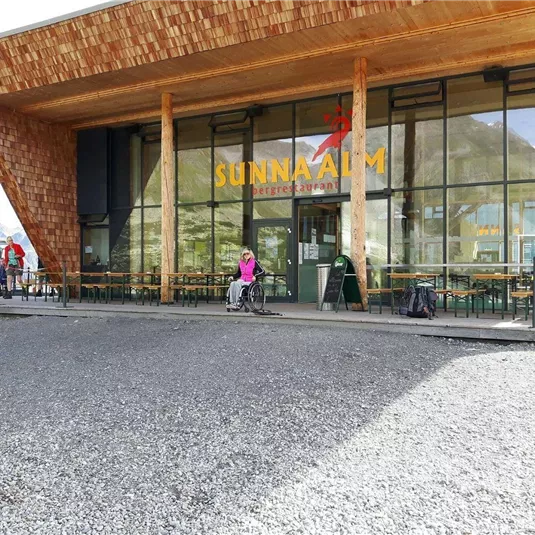 Ein modernes Restaurant mit großen Fenstern und einer Holzfassade in den Bergen. Vor dem Gebäude stehen einige Gäste und Tische im Freien.