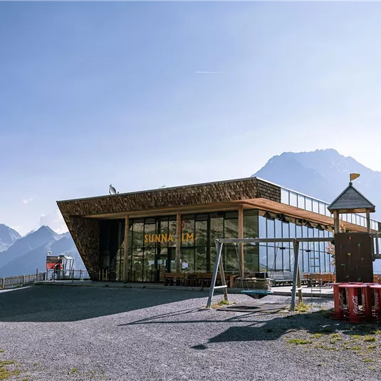 A modern building in the mountains with large windows and a view of the landscape. The sky is clear and the surroundings are green and mountainous.