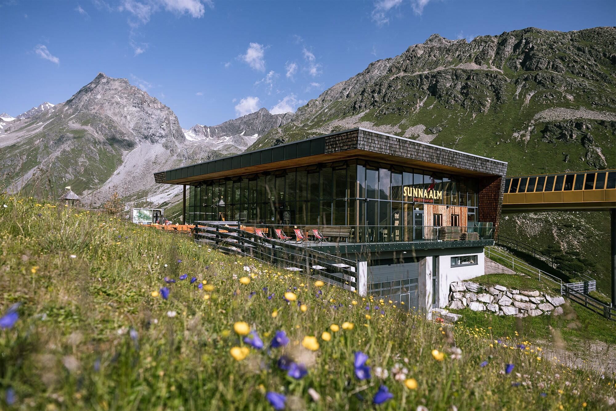 Ein modernes Gebäude in den Bergen, umgeben von blühenden Wiesen und majestätischen Felsen. Die Aussicht ist klar und sonnig, ideal für Erholung in der Natur.
