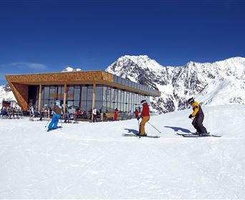 Eine moderne Berghütte inmitten schneebedeckter Berge. Skifahrer genießen den Tag bei klarem Himmel und Sonnenschein.