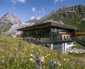 A modern building in a mountainous landscape with blooming meadows. In the background, there are majestic mountains and a blue sky.
