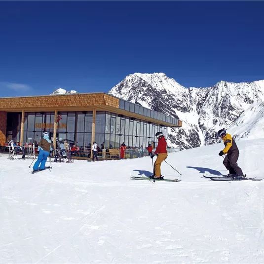 Eine moderne Berghütte umgeben von schneebedeckten Bergen. Skifahrer genießen die Sonne auf einer Piste.