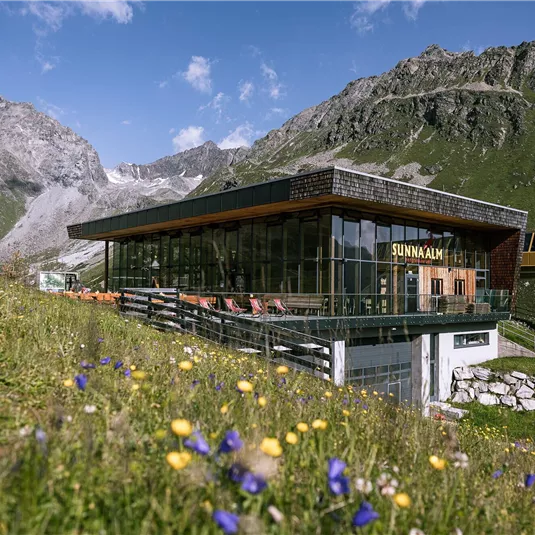 A modern building in a mountainous landscape with blooming meadows. In the background, there are majestic mountains and a blue sky.