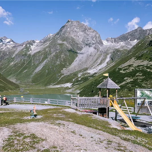 Ein Spielplatz in den Bergen mit einer Rutsche und einer Schaukelturm. Im Hintergrund sind grüne Hügel und hohe Berge zu sehen.